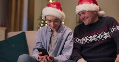 Dedenle kanepede oturan ve telefonu tutan neşeli genç adam. İnternette bulduğu komik videoları gösteriyor. Büyükbaba telefona bakar ve samimi bir şekilde gülümser. Noel Baba şapkası giymişler..