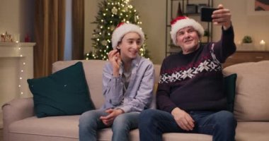 Torun ve büyükbaba oturma odasında kanepede oturuyorlar. Telefonda akrabalarıyla görüntülü görüşme yapıyorlar, konuşuyorlar, gülümsüyorlar. Noel Baba şapkası takıyorlar. Odada bir Noel ağacı sallanıyor..