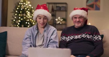 Noel arifesi. Oğlu ve babası kanepede oturuyor ve akrabalarıyla birlikte laptopta görüntülü görüşme yapıyorlar. İletişim kuruyorlar, gülümsüyorlar ve Noel şapkası takıyorlar. Arkasında titreyen bir Noel ağacı.