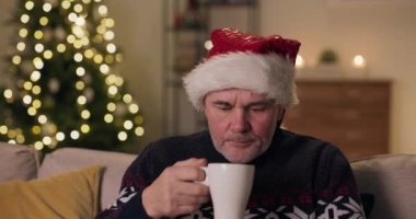 Kanepede oturan ve Noel Baba şapkası takan orta yaşlı bir adamın yakın çekimi. Adam bir fincan çay aldı ve bir yudum aldı. Sonra kameraya baktı. Noel 'de yalnız kaldığı için üzgün..