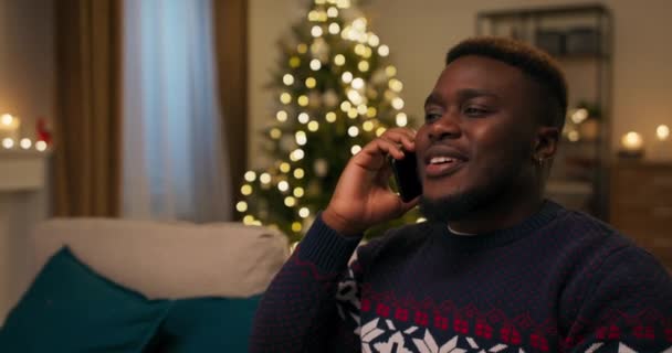 一个长相像非洲人的男人正在和女朋友通电话 他们正在计划圣诞节假期的旅行 这家伙心情很好 面带微笑 他穿着暖和的毛衣 耳朵里有球 — 图库视频影像