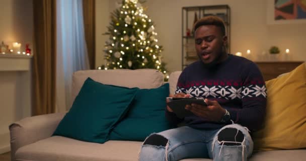 冬夜一个长相像非洲人的年轻人坐在沙发上 拿着石碑 他一边在社交媒体上翻阅新闻 一边笑着 房间里闪烁着一棵圣诞树 — 图库视频影像