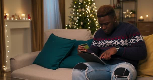 一个长相像非洲人的男人坐在沙发上 用笔记本电脑在网上商店里购物 他仔细地输入卡片的细节 并支付购买费用 房间里装饰一新 圣诞树闪烁着光芒 — 图库视频影像