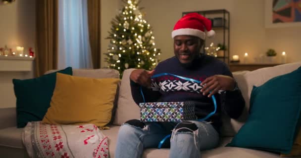 平安夜一个看起来像非洲人的家伙坐在沙发上 戴着圣诞老人的帽子 打开礼物 他打开盒子 看到了他不需要的温暖的耳机 男孩心烦意乱地把它们放在一边 — 图库视频影像