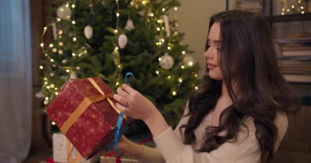 友人のための長い髪パックの贈り物と魅力的なブルネット 女の子は青いリボンの弓を結び それに合った箱を選択します 輝くクリスマスツリーの後ろで 美しさは笑っています — ストック動画
