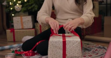 Arkadaşlar için hediye paketleyen kızların yakın çekimleri. Esmer, kırmızı kurdeleyi pruvaya bağlar, makası alır ve keser. Işıldayan Noel ağacının ve hediyelerin arkasında.