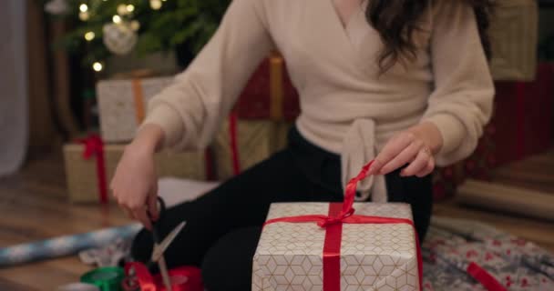 年轻的黑发女孩的特写 长发包裹着圣诞礼物 女孩用蝴蝶结把红丝带绑在盒子上 然后割断了丝带 在闪闪发光的圣诞树和礼物后面 — 图库视频影像