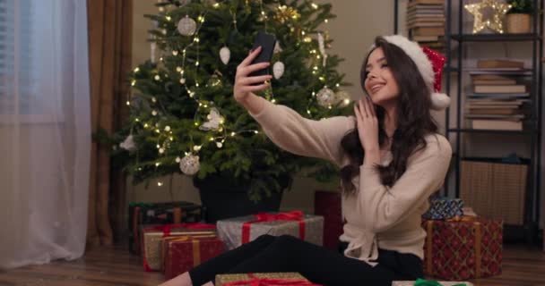 長い髪と頭の上にサンタクロースの帽子を持つ魅力的なブルネットはクリスマスツリーの近くの床に座って自撮りをします 女の子はポーズをとって写真を撮り 笑顔で友人に写真を送ります — ストック動画