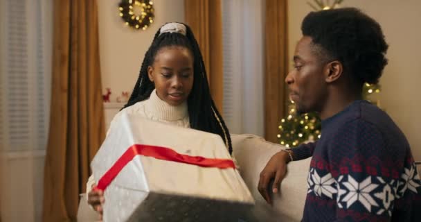 闪亮的圣诞树 两个长相像非洲人的年轻人坐在客厅的沙发上聊天 女孩把给哥哥准备的礼物手牵着 用红带捆起来 — 图库视频影像