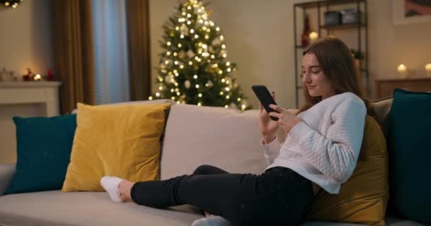 冬夜下班后 女孩坐在客厅的沙发上 美女手里拿着电话 看着滑稽的照片 微笑着 房间里很舒适 闪闪发光的圣诞树 — 图库视频影像