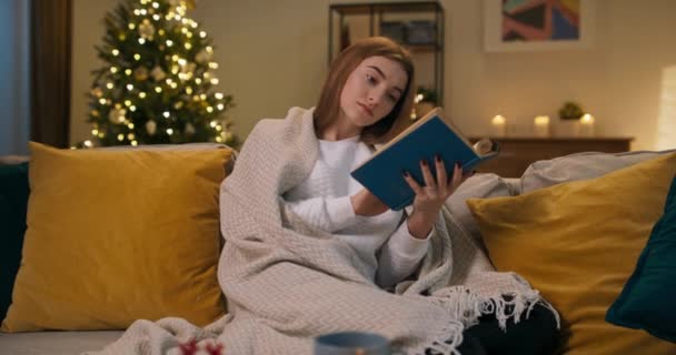 一个孤独的女孩坐在客厅的沙发上 她浑身毛毯 手里拿着一本蓝皮书 这个女孩读小说 反思自己所读的东西 房间里闪烁着一棵圣诞树 — 图库视频影像
