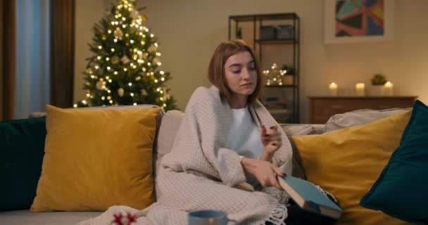 一个舒适的冬夜 一个棕色头发的女孩坐在客厅的沙发上看书 她把书合上 用毯子把自己包裹起来 沉思着自己所读到的东西 一棵圣诞树摇曳着 — 图库视频影像