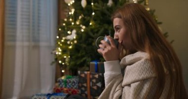 Noel ağacında çay içen gülümseyen güzel bir kızın portresi ışıklarla aydınlandı. Kadın battaniyeye sarılı sıcak içecekle kendini ısıtıyor..