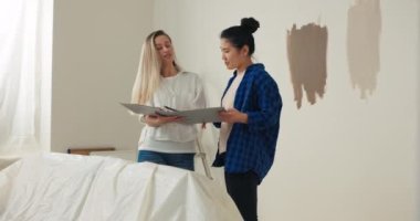 İki genç, gülümseyen kadın tadilat altındaki yeni bir dairede duruyorlar. Sarışın bir kadın, elinde daha fazla onarım planı olan bir dosya tutuyor. Başka bir kadın gelecek duvarların rengini seçiyor..