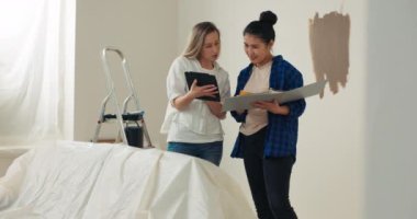 Tadilatın yapıldığı odanın ortasında büyük bir kanepe var ve arkasında iki genç kadın, bir merdiven, bir elektrik süpürgesi, resimler, kutular, boya silindirleri vs..