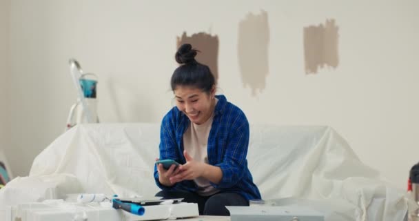 一个女孩坐在一个翻新过的公寓里的沙发上 手里拿着电话 这名妇女获得了有关快速交付选定家具的信息 — 图库视频影像