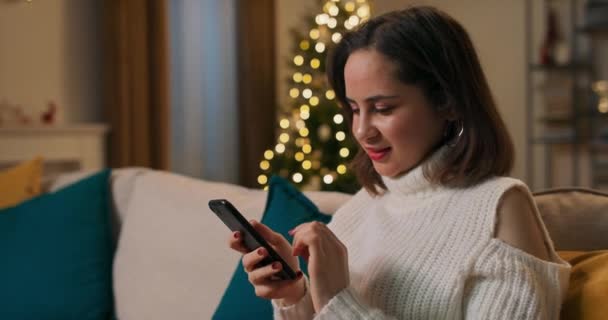 一个年轻的学生正在她的宿舍里过圣诞节 女孩坐在沙发上 点着圣诞树灯 向朋友和家人问好 — 图库视频影像