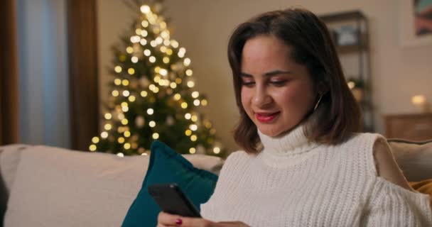 一个微笑的女孩一个人在公寓里过圣诞节 这个女人晚上坐在沙发上 点着圣诞树灯 在社交网络上和朋友们开玩笑地聊天 — 图库视频影像
