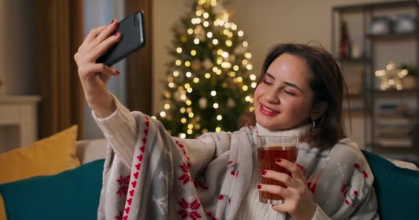 一个女孩穿着毛毯度过了一个孤独的冬夜 有影响力的人一边喝茶 一边在社交媒体上用手机自拍 背景是一株闪闪发光的圣诞树 — 图库视频影像