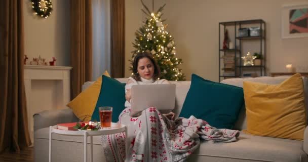 迷人的小女孩被毛毯盖住躺在沙发上 在圣诞节系列电脑上看电影 在圣诞树旁放松自己 — 图库视频影像