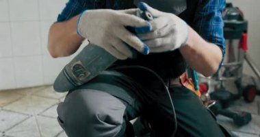 Bir evi kendi başına yenileyen genç bir adam yerde diz çöker iş kıyafetleri ile fayansların tahrip edilmesi için ekipman hazırlar, kablo çekiçle çekiç, inşaat tecrübesi.