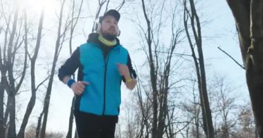 Sakallı genç adam sabah parkta koşar ve düzgün nefes alır. Mavi ve siyah spor giyimli ve başında şapka ve kulaklık var. Adamın bilekliği ve saati var..