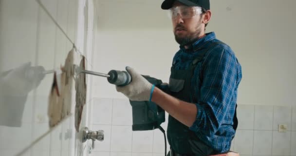 シャツと帽子を身に着けている作業服の男は 家の改装を行うキッチンバスルームでタイルをハンマー — ストック動画