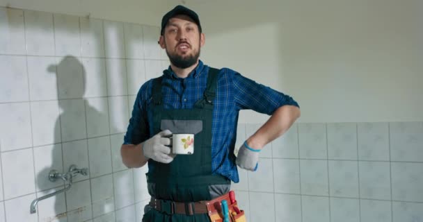 戸建住宅の改修工事に従事する忙しい労働者 仕事中の男は野球帽のリラックスが目を覚ますために窓のシップコーヒーを見ています カメラを見ると — ストック動画