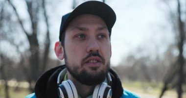 Kulaklık takan, kameraya bakıp gülümseyen sakallı genç adamın yakın çekimi. Başında siyah şapkası, elinde saati ve parmağında alyansı var. Adam spor kıyafetleri giymiş..