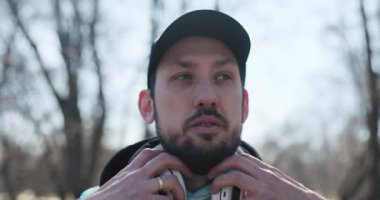 Kulaklık takan, kameraya bakıp gülümseyen sakallı genç adamın yakın çekimi. Başında siyah şapkası, elinde saati ve parmağında alyansı var. Adam spor kıyafetleri giymiş..