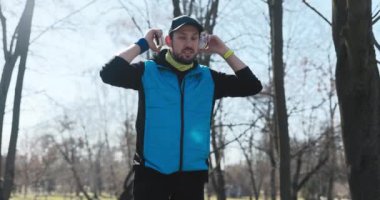 Sakallı adam parkta duruyor ve kulaklık takıyor. Oğlan mavi ve siyah elbiseler giymiş ve şapkası var. Elinde bir saat var. Kameraya bakar, ellerini birleştirir ve gülümser..