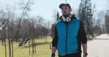 Genç sporcu parkta antrenmandan sonra yorgun bir şekilde yürür. Nefes verdi ve kaputu kafasına geçirdi. Adam mavi ve siyah giyinmiş ve şapka ve kulaklıkları var. Elinizde yeşil kayışlı bir saat var..