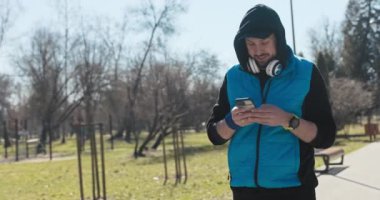 Sakallı adam karısına mesaj atıyor. Parkta yürüyor, telefonu elinde tutuyor ve gülümsüyor. Oğlan mavi ve siyah giyinmiş ve kafasında bir başlık var. Kulaklığı ve saati var..
