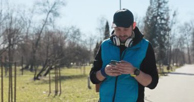 Spor kıyafetleri giymiş genç bir baba parkta elini kolunu sallayarak yürüyor. Adam mesaj atıyor, etrafa bakıyor ve gülümsüyor. Başında şapka ve kulaklığı var. Elinde saat var..