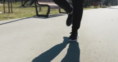 Parkta koşan adamın bacaklarına yakın çekim. Çocuk siyah pantolon ve beyaz tabanlı spor ayakkabı giyiyor. İnsan hızlı koşar, güneş parlar ve asfalta gölge yansıtır..