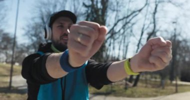 Egzersiz yapan insanların yakın çekimi. Adam parkta duruyor ve ellerini uzatıyor. Elinde saat, parmağında yüzük var. Kafasında müzik çalan bir şapka ve kulaklık var. Düzgün nefes alıyor.