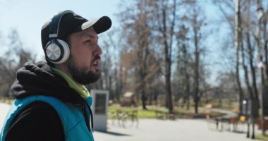 Güneşli bir gün. Adam parkta spor yapıyor. Durdu ve nefes verdi. Şapkalı ve kulaklıklı bir adam elinde bir şişe su tutuyor ve ondan içiyor. Elinde saat var ve siyah kapüşonlu giyiyor..