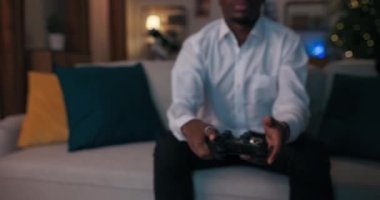 Tahta masanın üzerinde duran joystick 'in yakın çekimi. Afrikalı görünümlü bir adam kanepeye oturur, joystick alır ve video oyunları oynamaya başlar. O, beyaz gömlek giyiyor. Öğrenci oyuna odaklanmış..