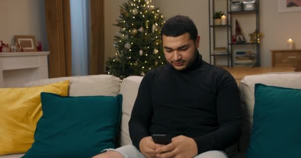 迷人的黑发女人坐在客厅的沙发上 他穿着黑色毛衣 拿着电话 一个男人和朋友聊天 一棵圣诞树在身后摇曳 — 图库视频影像
