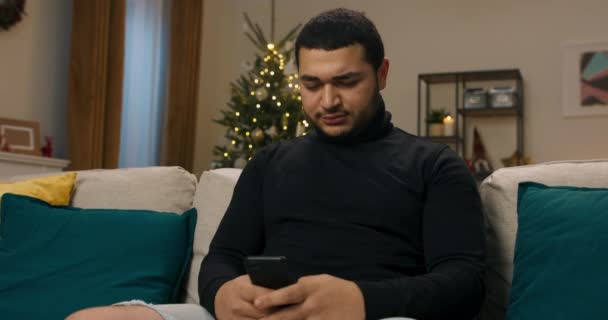 一个黑头发的年轻人穿着黑色毛衣坐在沙发上 圣诞树在后面闪烁着 他手里拿着电话 专心致志地打字 然后他看着相机笑了 — 图库视频影像