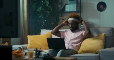 Afrikalı görünümlü adam kanepede oturuyor ve dizüstü bilgisayar tutuyor. Kulaklık takar ve iş arkadaşlarıyla görüntülü konuşmaya başlar. Adam merhaba dedi ve iletişim kuruyor. Kanepede sarı yastıklar var..