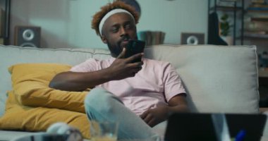 Kıvırcık saçlı Afrikalı öğrenci koltukta oturuyor, sarı yastıklara yaslanıyor ve telefonu tutuyor. Kız arkadaşı onu aradı. Çocuk gülümsedi ve telefonu açtı. İletişim kurduğu için mutlu..