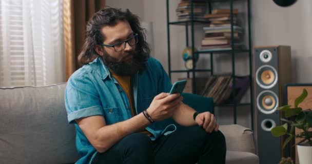 一个留着胡子的英俊男人和他的狗坐在客厅的沙发上 这个满脸胡子的男人正在智能手机上浏览保险单的报价 — 图库视频影像