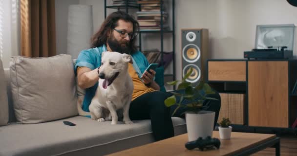 一个留着胡子的男人在狗的陪伴下 用智能手机浏览网络 一个心满意足的小男孩坐在客厅沙发上 一边微笑着一边抚摸着宠物 一边用手机 — 图库视频影像