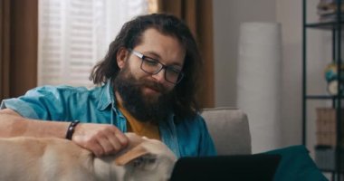 Sevimli, krema renkli bir köpek, sahibine dizüstü bilgisayara belge yazması için eşlik ediyor. Hayvan, evdeki kanepede uzaktan çalışan adamın yanında bilgisayara bakar..