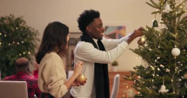 Gözlüklü Asyalı kız ve süveter ve siyah eşarplı Afrikalı oğlan Noel ağacının yanında duruyor. Süslemeleri ellerinde tutuyorlar ve ağaca tutunuyorlar. Arkadaşlar gülümsüyor..