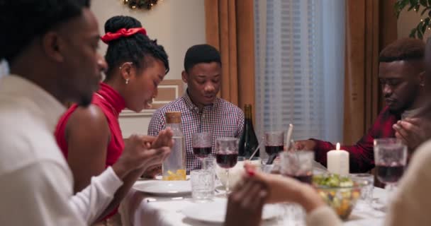 クリスマスには アフリカ系の友達がお祝いの席に座りました 彼らは手を取り 目を閉じて祈ります シャツの男が祈りを言った 学生たちは乾杯し 皆でワインを飲みました — ストック動画