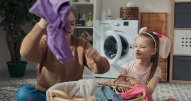 Genç anne, küçük kızına kıyafetleri renklerine göre doğru sıralamayı öğretiyor. Mor pantolonu sepetten çıkardı, katladı ve kenara koydu. Kız dikkatle izliyor. Konuşuyorlar ve gülümsüyorlar..