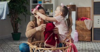 Anne ve kızı banyoda yerde oturuyor ve giysilerini katlıyorlar. Giysileri sepetten çıkarıyorlar, gülüyorlar ve oynamaya başlıyorlar, kıyafetlerini birbirlerinin kafalarına geçiriyorlar. Çamaşır makinesi var..