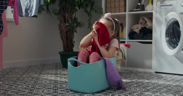6岁的长发女孩坐在蓝色的洗碗里 她把洗干净的衣服抽出来闻 闻起来又香又新鲜 洗好的衣服挂在旁边 女孩玩得很开心 — 图库视频影像